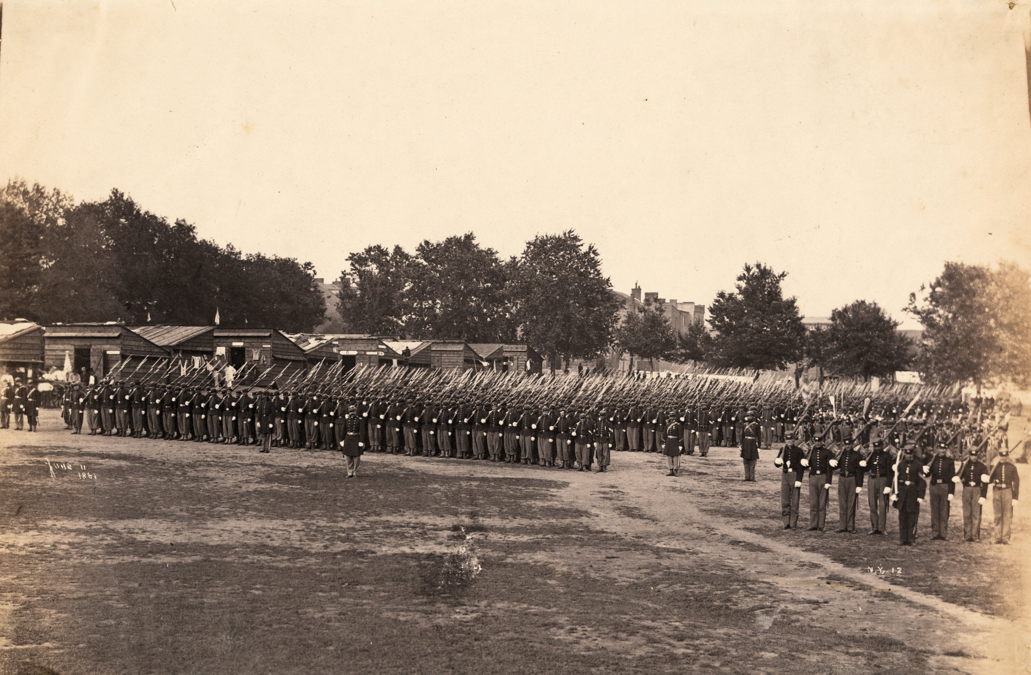 Photograph showing Civil War Union soldiers and Zouaves in formation at Franklin Square, Washington, D.C.; small rough buildings, probably barracks, in background